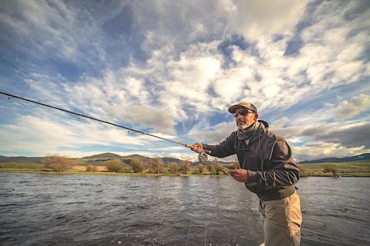 Person wearing hat and sunglasses casting fishing line into river under cloudy blue sky