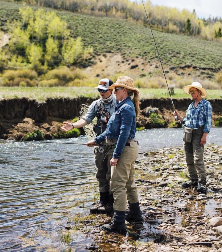 Two people standing by a river wearing hats and sunglasses one holding a fishing rod