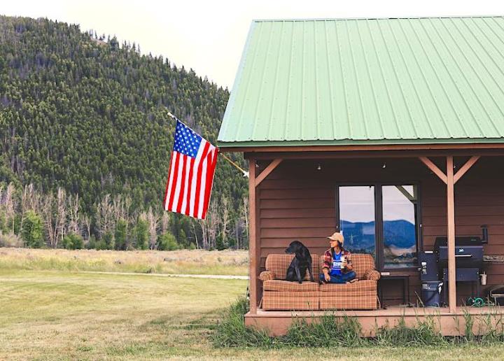 Person and brown dog sitting on sofa on porch of wooden cabin with a green roof and US flag